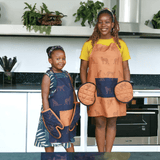 A woman wearing an amber-colored kitchen apron with a repeating cheetah print and a large navy blue pocket at the center front for a striking and functional design and circular pot holders on her hands standing next to a child wearing navy blue kitchen apron adorned with a cheetah pattern, featuring a contrasting amber-colored central pocket offering a blend of functionality and playful style with traditional oven mits in navy.