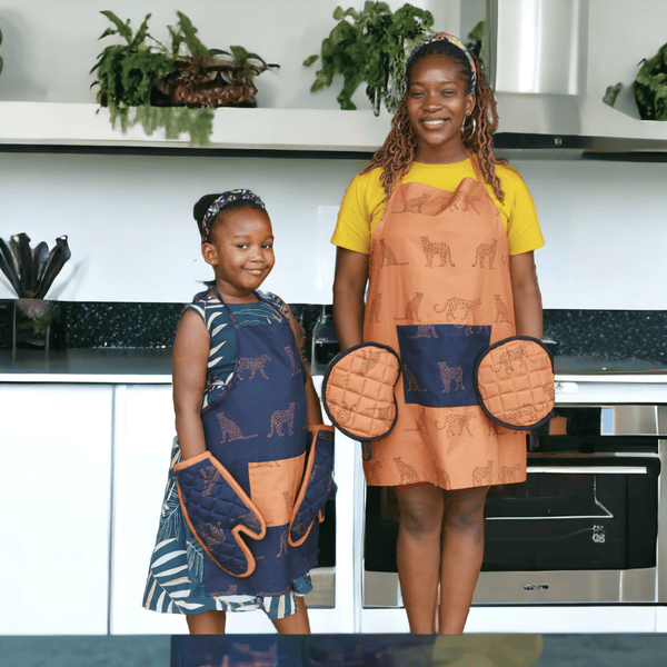 A woman wearing an amber-colored kitchen apron with a repeating cheetah print and a large navy blue pocket at the center front for a striking and functional design and circular pot holders on her hands standing next to a child wearing navy blue kitchen apron adorned with a cheetah pattern, featuring a contrasting amber-colored central pocket offering a blend of functionality and playful style with traditional oven mits in navy.