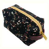 Floral-patterned cosmetic pouch with a purple logo patch and zipper, featuring a black base with red, yellow, and white accents, side view