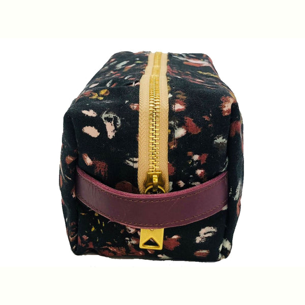 Floral-patterned cosmetic pouch with a purple logo patch and zipper, featuring a black base with red, yellow, and white accents, end view